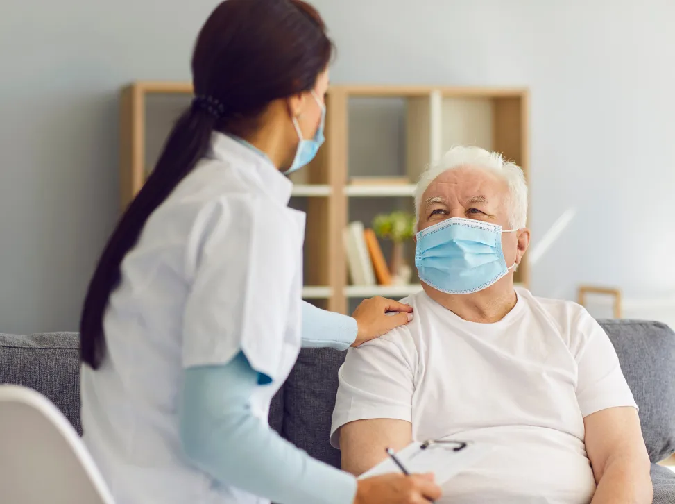 Healthcare worker in scrubs and mask comforting an elderly man in a mask, seated on a couch in a room.