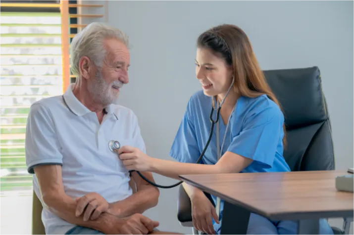 chronic-1 A nurse smiles while using a stethoscope to check an older man's heartbeat in a medical office.