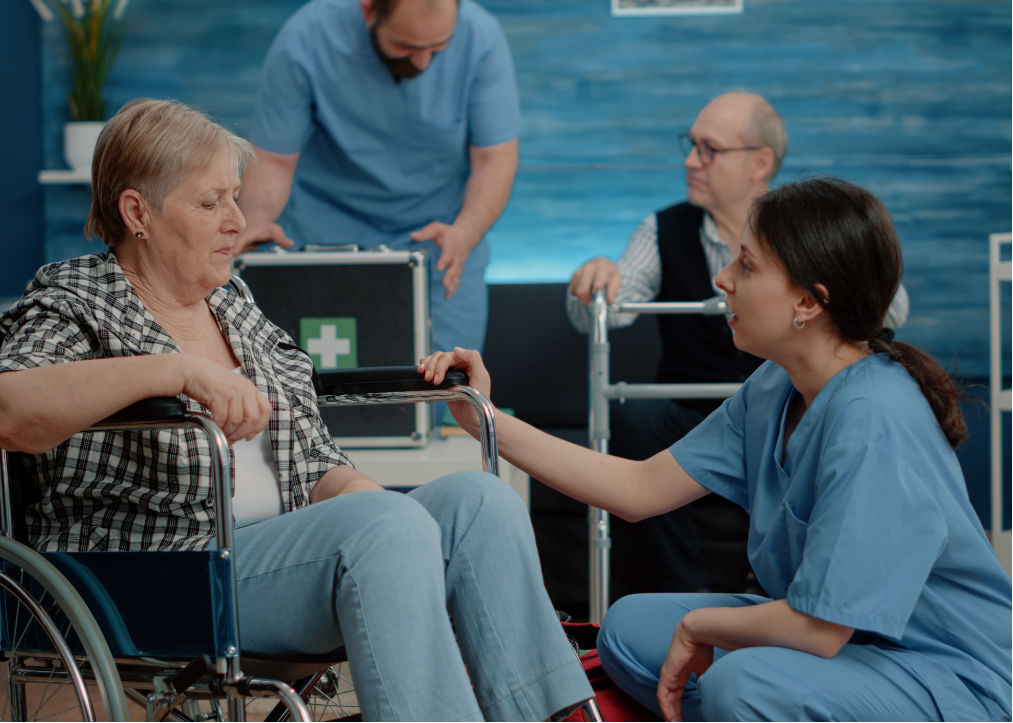 chronic-disease-management-1 A nurse crouches beside an elderly woman in a wheelchair, with two paramedics working in the background.