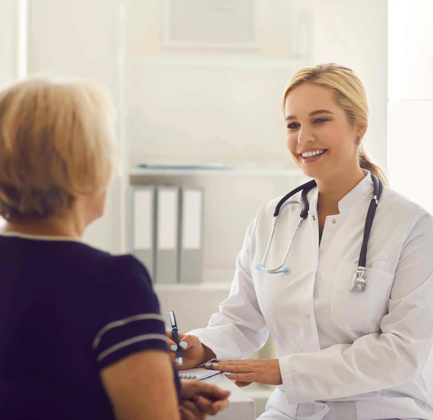 A smiling female doctor with a stethoscope talks to an older female patient in a medical office.