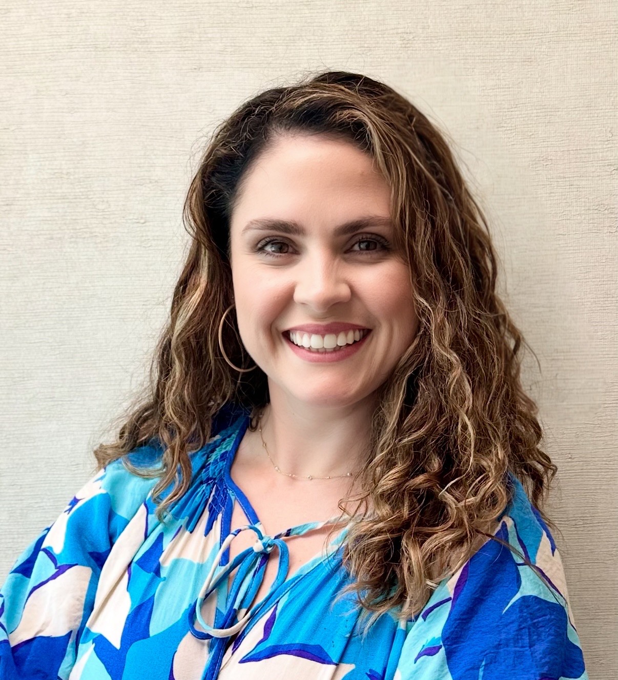 HANNAH SWINDELL Woman with curly brown hair and blue patterned blouse smiling in front of a plain light background.