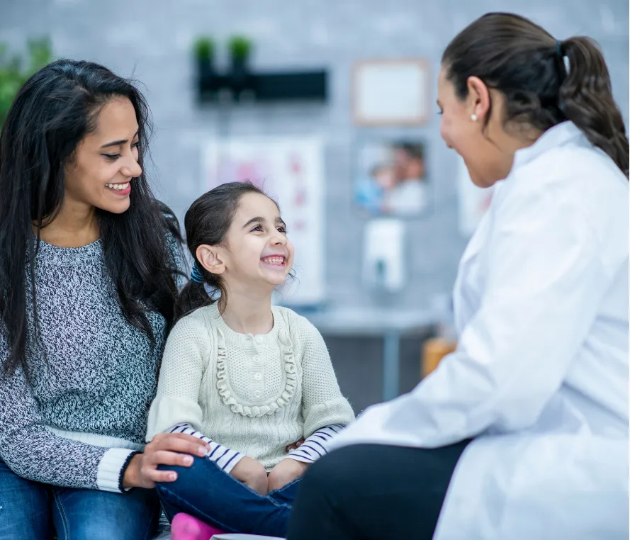about-4 A doctor smiles and talks with a mother and her smiling young daughter in a medical office.