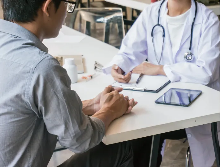 A patient talks to a doctor with a stethoscope, clipboard, and tablet on the desk between them.