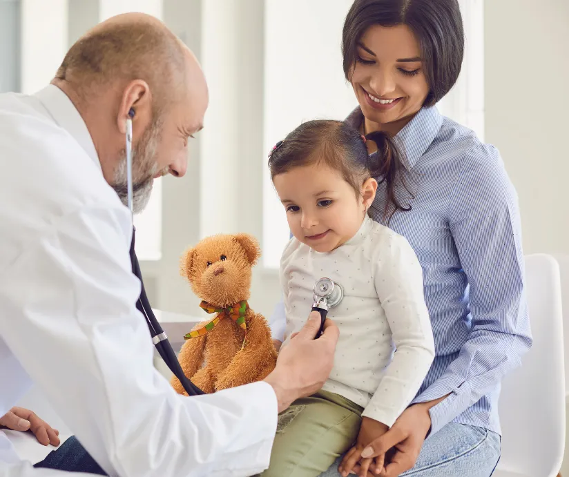 Doctor uses stethoscope on young girl sitting on her mother’s lap, with a teddy bear nearby.