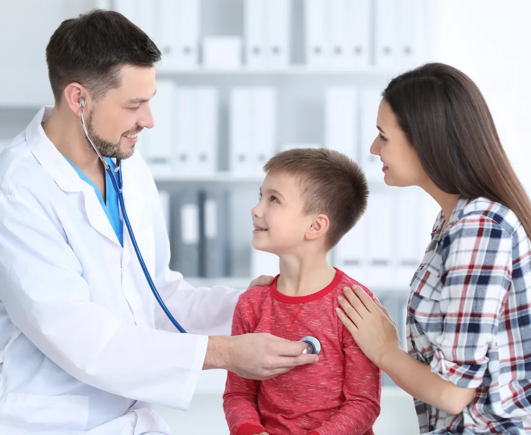 A doctor uses a stethoscope on a smiling boy as his mother sits beside him, gently touching his shoulder.