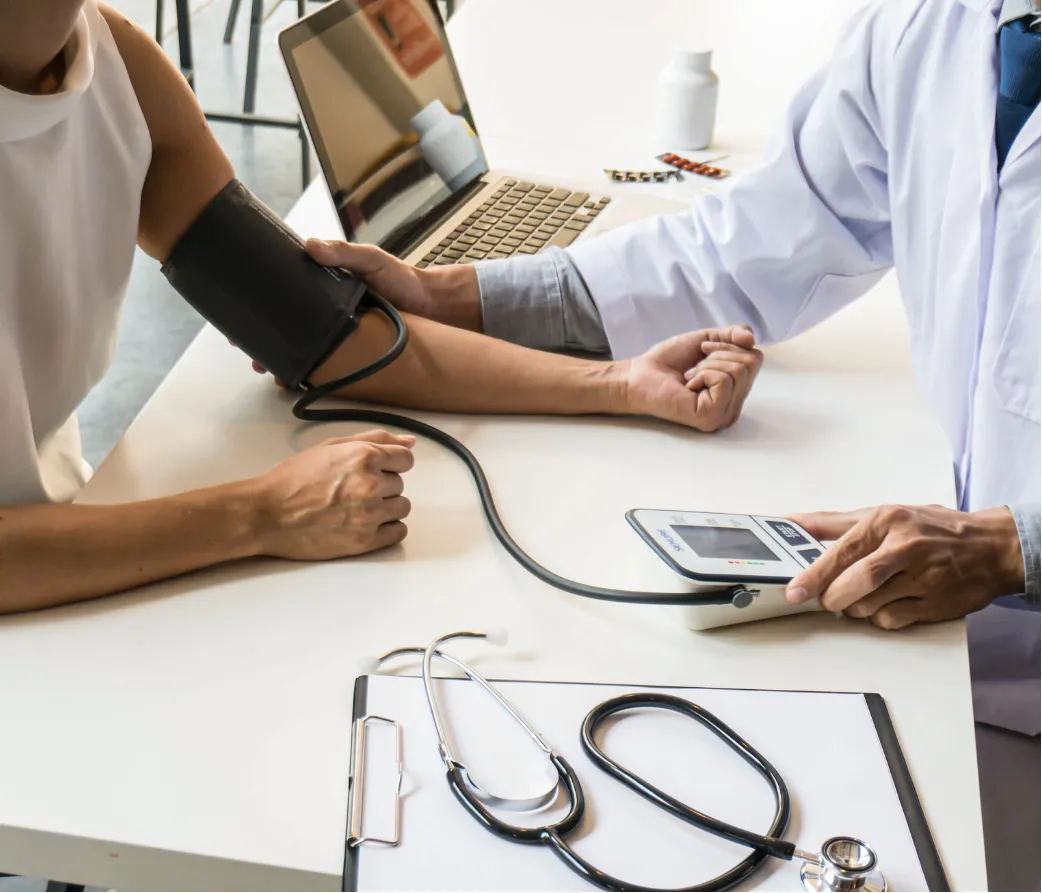 A doctor checks a patient's blood pressure at a desk with a laptop, stethoscope, and medicines.